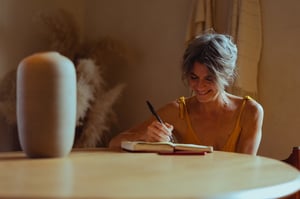 older woman sitting at table smiling while writing in journal