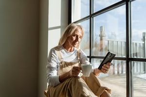 older woman sitting in the window at home drinking coffee with smartphone