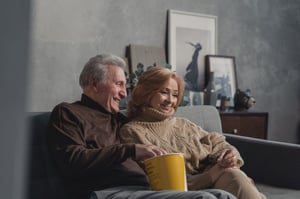 older couple in warm clothers sweaters sitting on couch watching a movie with popcorn